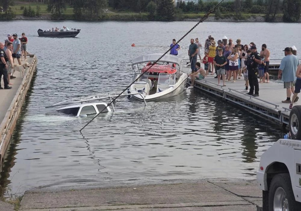 A partially submerged car is in the water next to a dock, with a boat nearby. A crowd of people stands on the dock watching, and another boat is seen in the background.