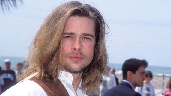 A person with long blond hair and a short beard wearing a white shirt stands outdoors, with blurred people and a beach in the background.