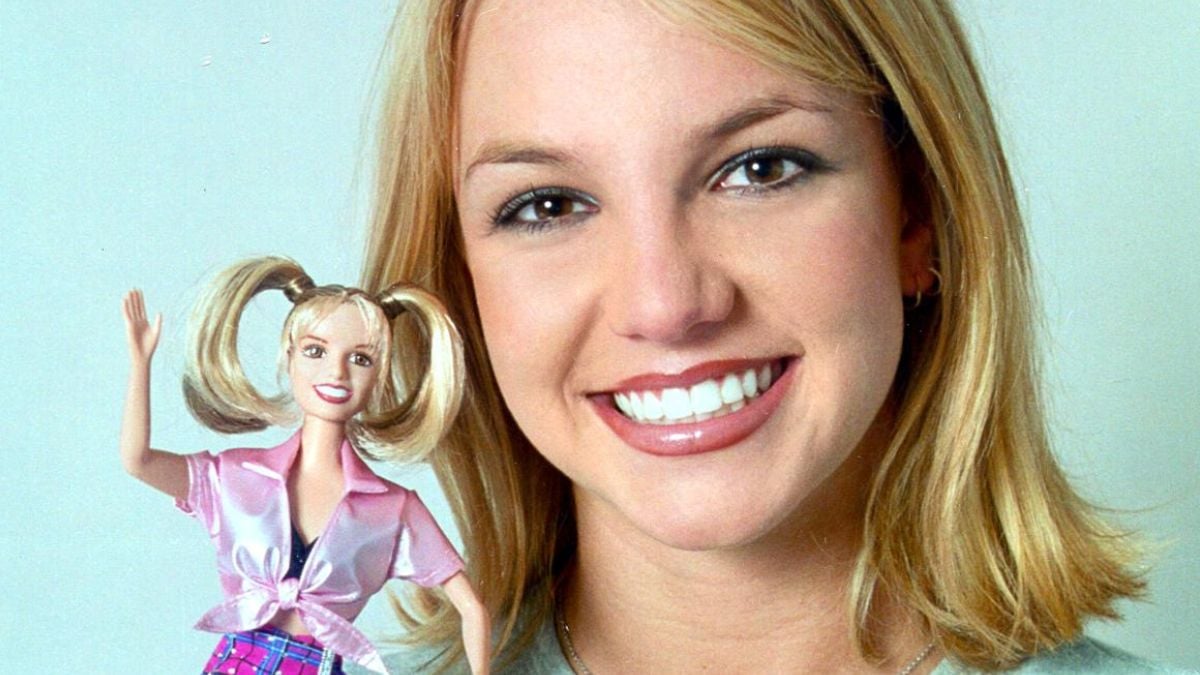 A smiling woman with blonde hair holds a doll that resembles her, also with blonde hair and wearing a pink outfit. Both are facing the camera against a plain light background.