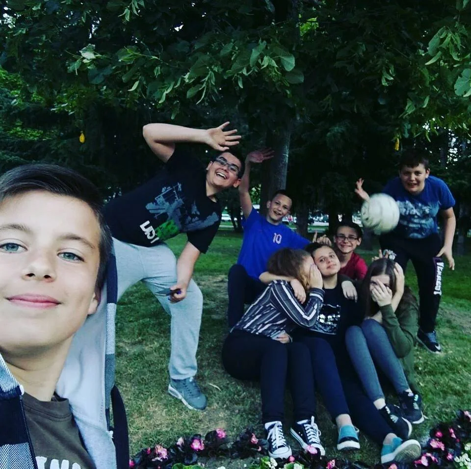 A group of kids pose and smile under a tree in a park. One boy takes a selfie while others wave and laugh, sitting or crouching together.
