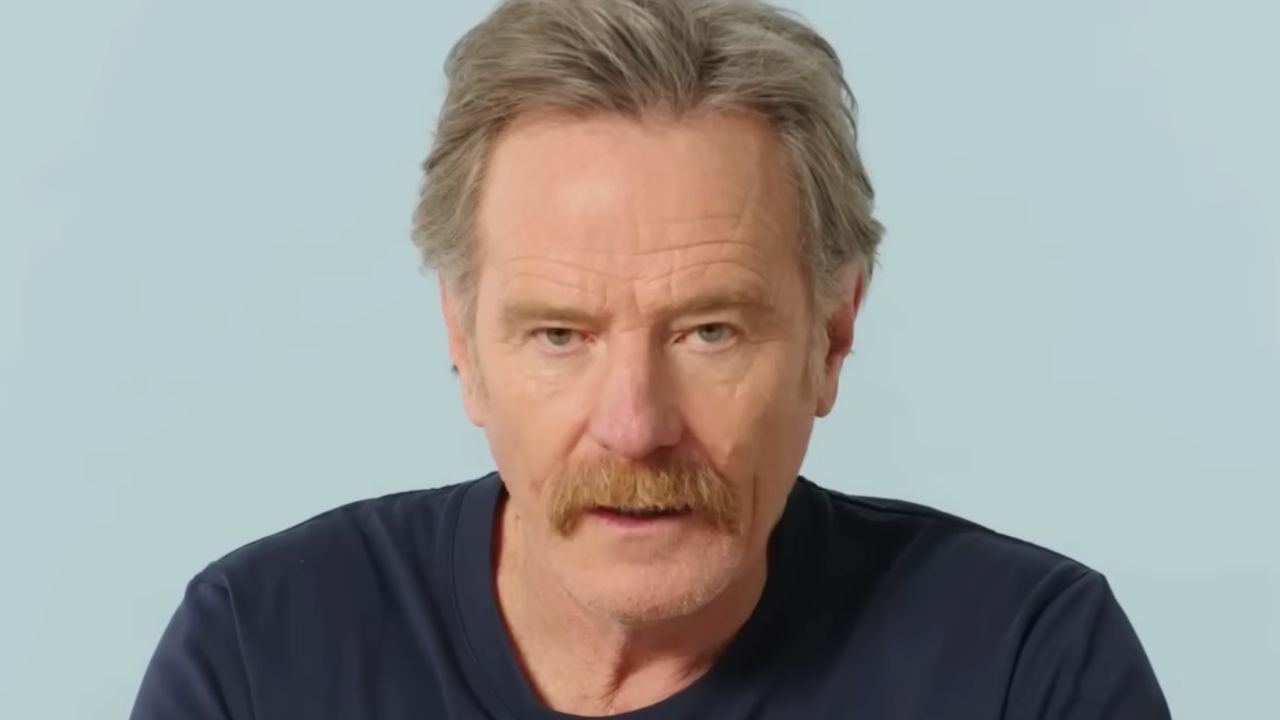 A middle-aged man with light skin, gray hair, and a mustache, wearing a dark shirt, looks directly at the camera against a plain light blue background.