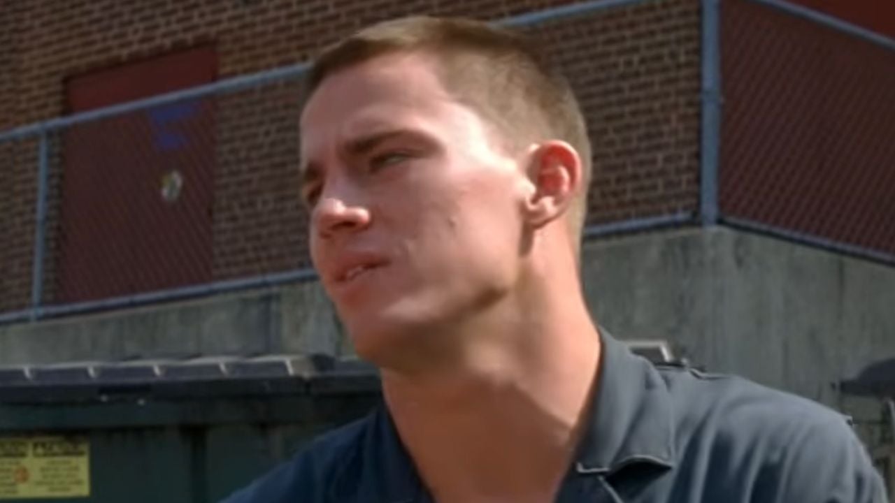 A man with short hair in a dark shirt stands outdoors in front of a brick wall and chain-link fence, looking to the side.