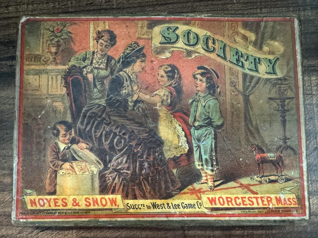 A vintage game box titled “Society” shows a woman and two children talking, while another child looks through a box. The box also reads “Noyes & Snow, Worcester, Mass.” with ornate Victorian-style art.