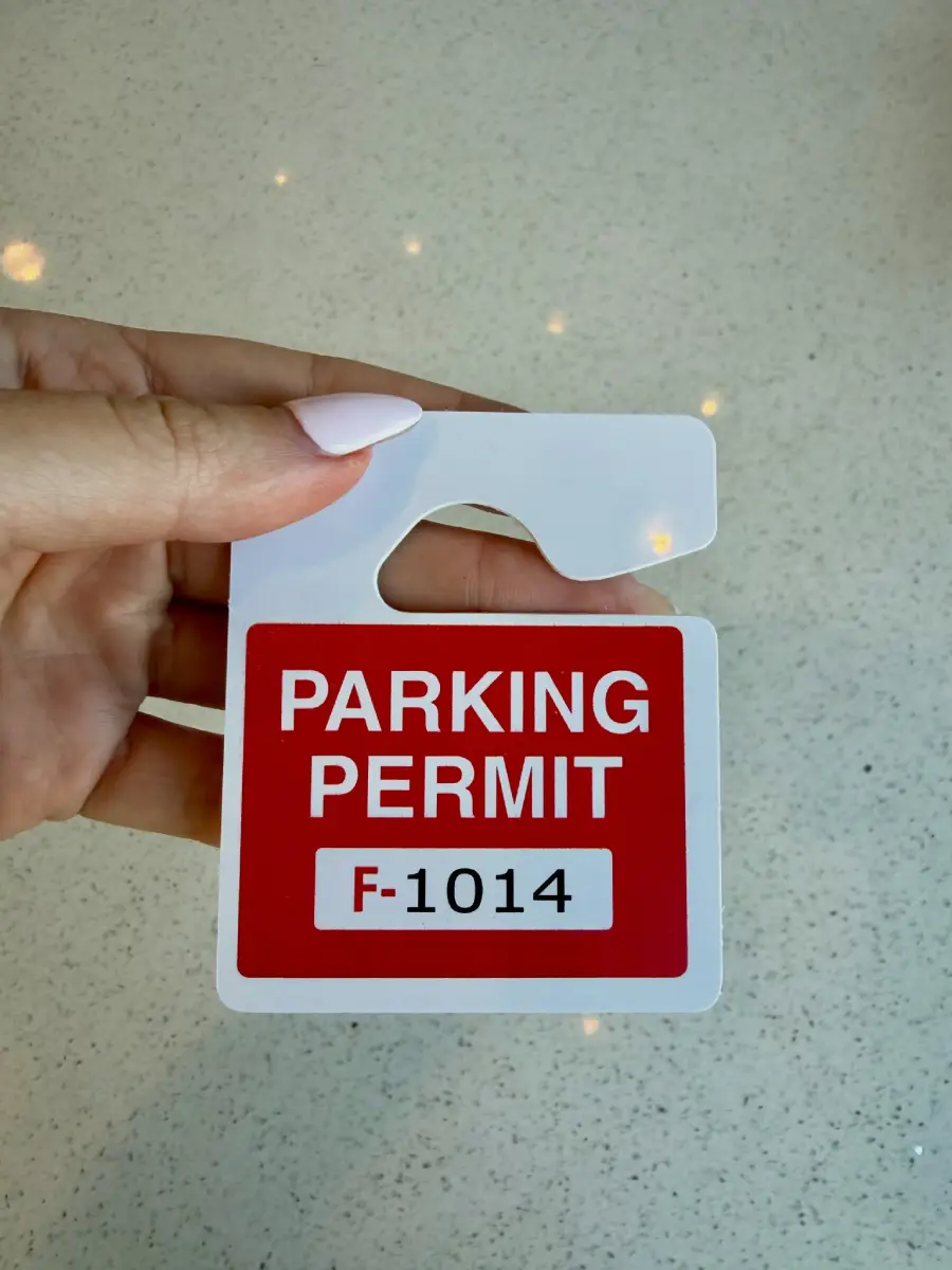 A hand with light pink nails holds a red and white parking permit labeled “PARKING PERMIT F-1014” against a speckled light countertop background.