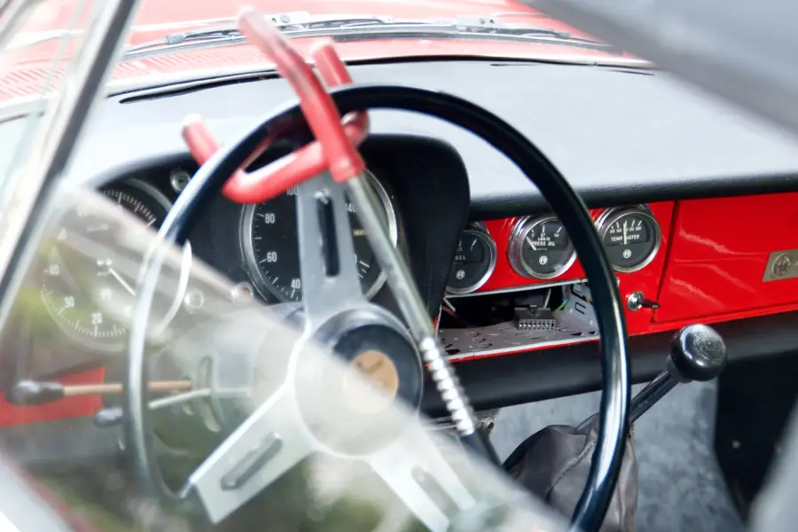 Close-up of a vintage car’s interior showing a black steering wheel, red dashboard with gauges, and a large red clamp attached to the steering wheel, possibly as a security device.