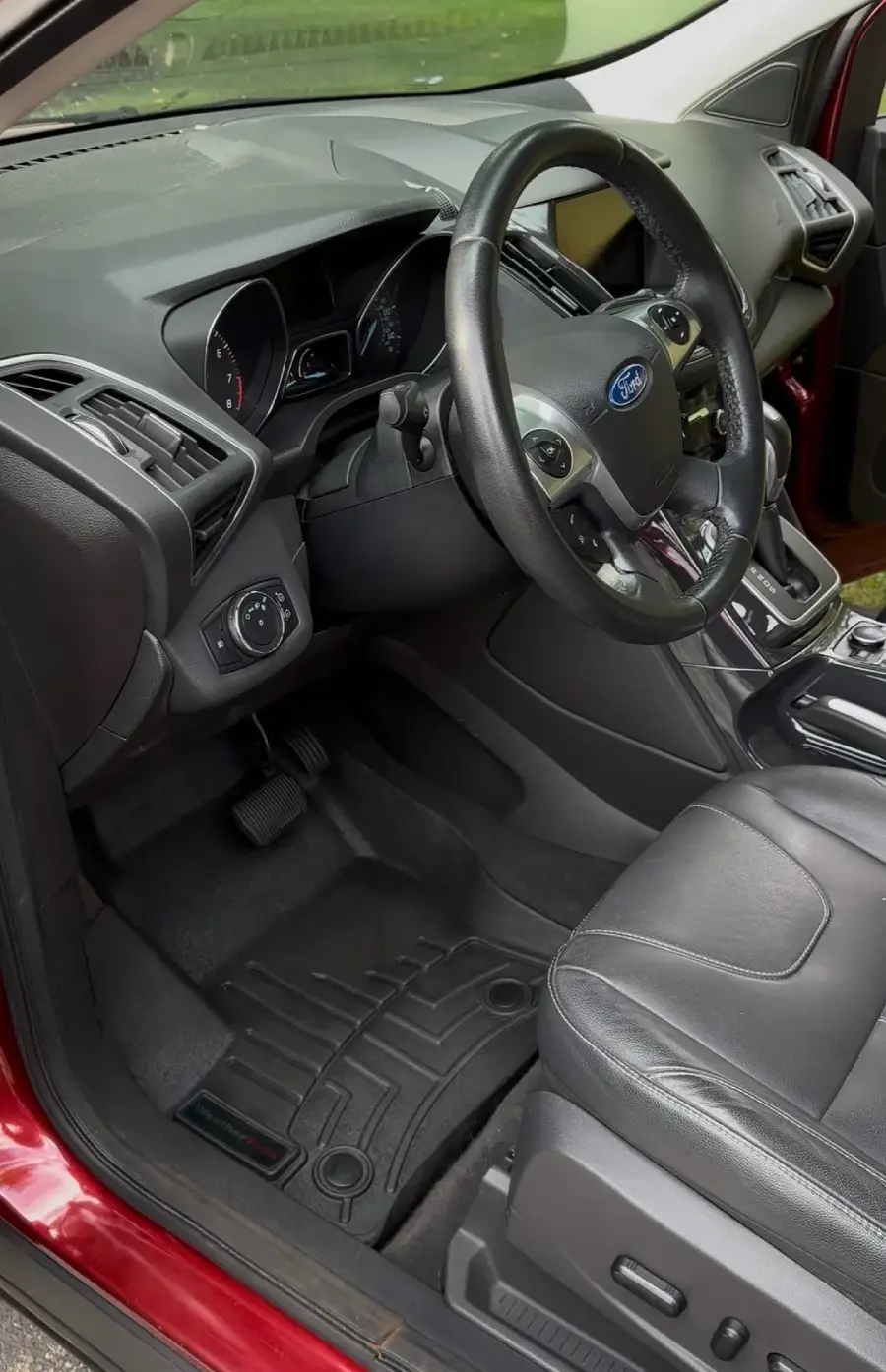 Driver’s side interior of a Ford vehicle showing the steering wheel, dashboard, pedals, and black leather seat. The door is open, revealing car controls and the center console.
