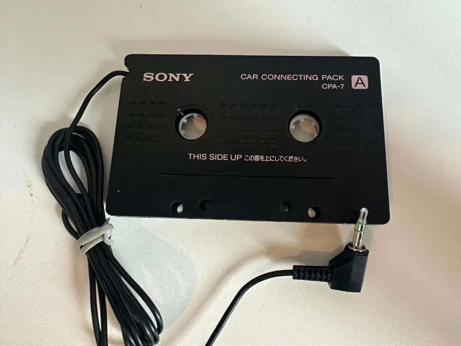 A black Sony car connecting cassette adapter with an attached audio cable and 3.5mm plug, placed on a white surface. The cassette is labeled “CAR CONNECTING PACK CPA-7” and “THIS SIDE UP.”