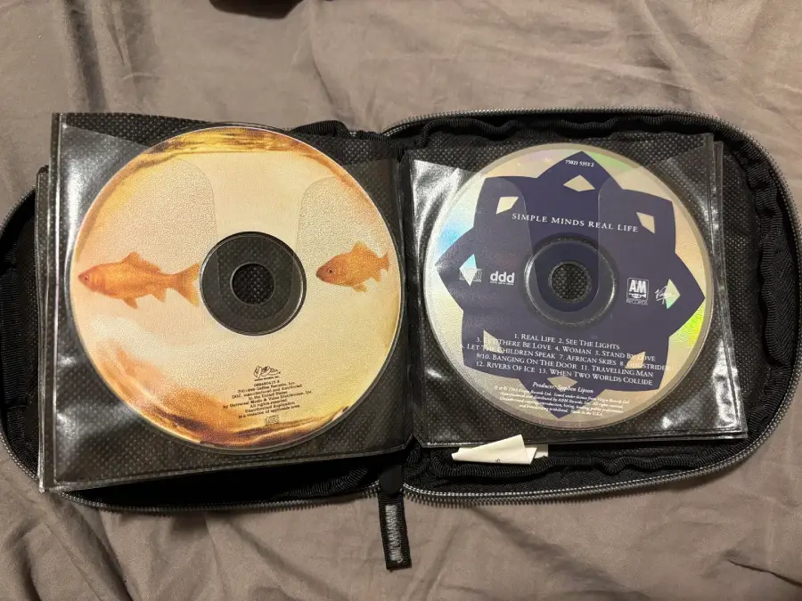 Two CDs in a black zippered case; the left CD features artwork with two goldfish, and the right CD is labeled “Simple Minds Real Life” with a blue and white geometric design.