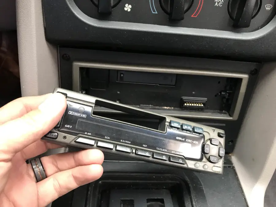 A hand holds a detached car stereo faceplate in front of a car dashboard, showing the empty stereo slot with visible connector pins.