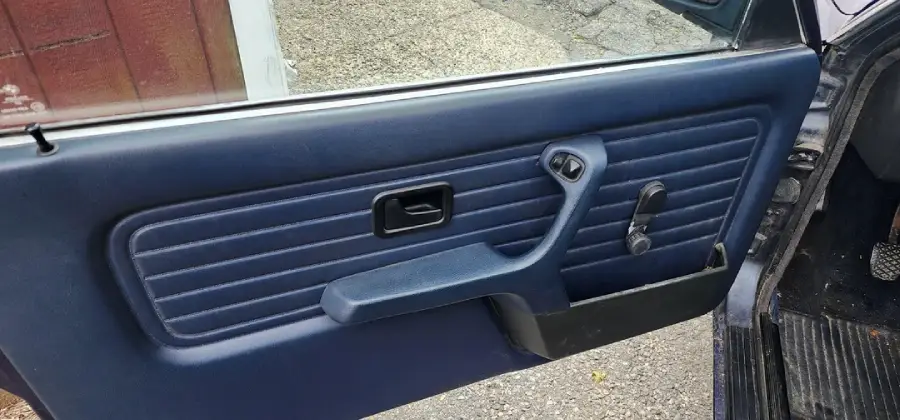 Blue car door interior with horizontal stitching, a black rectangular door handle, manual window crank, and an armrest. Car is parked on a paved surface.