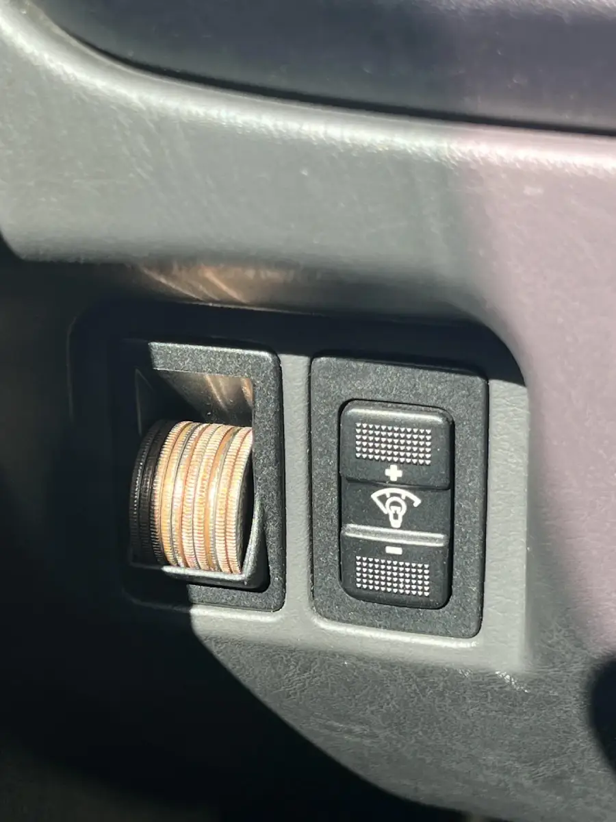 A stack of coins is stored in a circular compartment next to a pair of vehicle dashboard buttons, one with a windshield wiper symbol. Sunlight partially illuminates the area.
