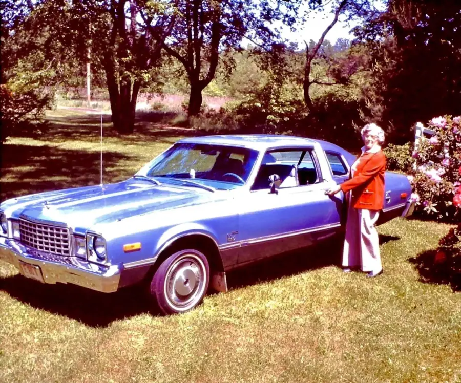 An older woman in a red jacket stands next to a vintage blue car parked on grass, with trees and bushes in the background on a sunny day.