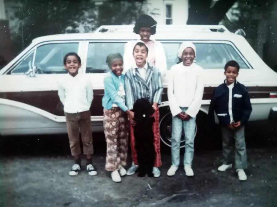 Six smiling children and one adult stand in front of a white station wagon. The children are dressed in 1970s-style clothing, and a small black dog is at their feet. The background includes trees and a house.