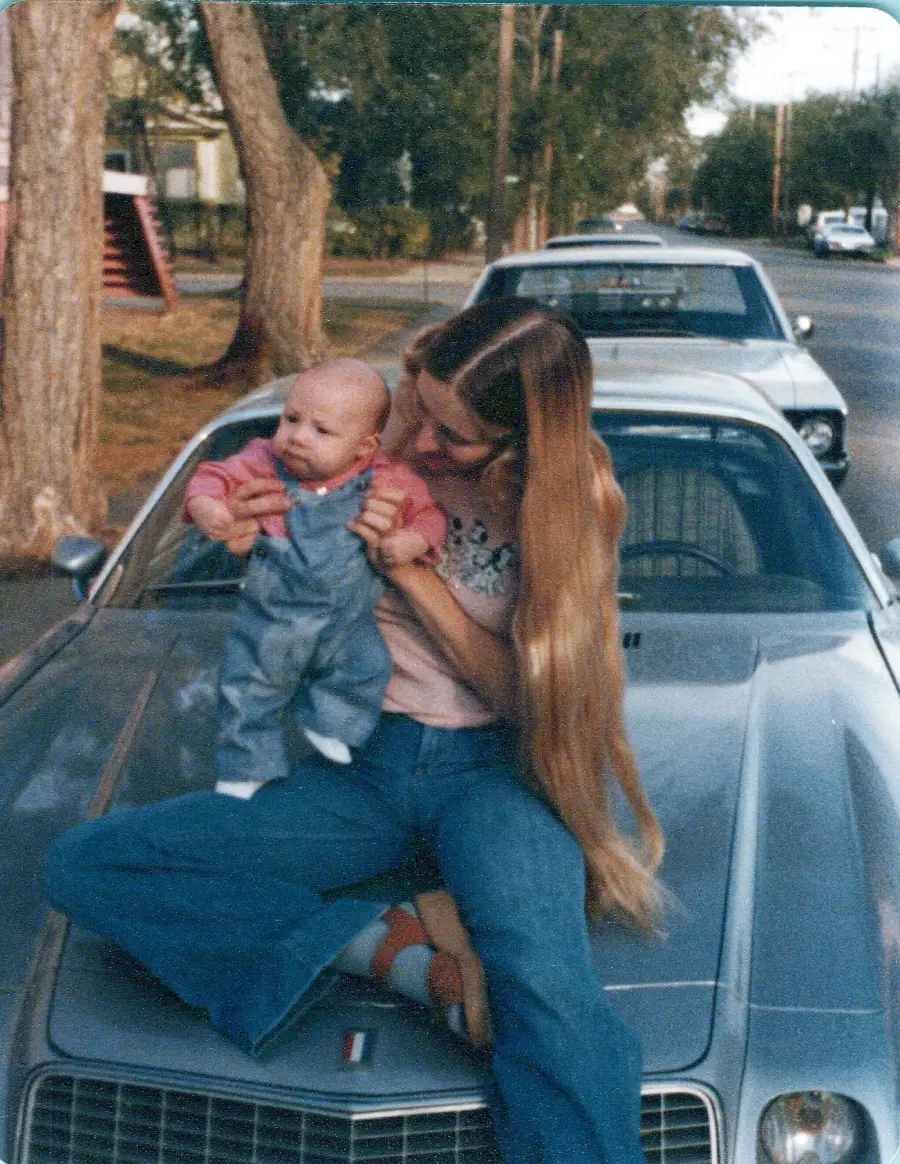A woman with long hair sits on a vintage car holding a baby in overalls, on a tree-lined street with parked cars in the background.