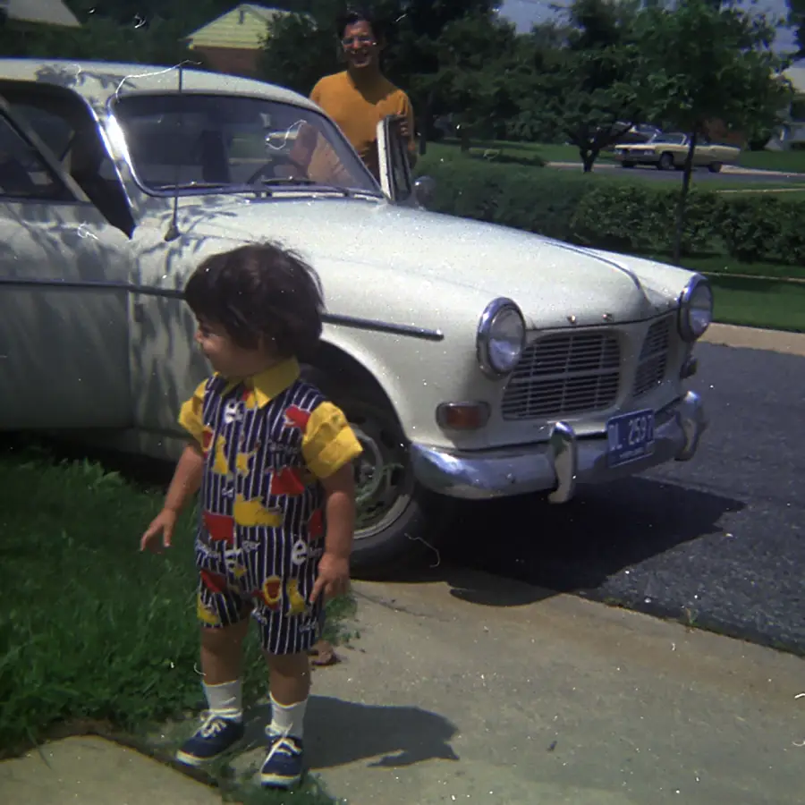 A young child in colorful clothes stands on a sidewalk near an old white car, with an adult smiling nearby. It’s a sunny day in a suburban neighborhood.