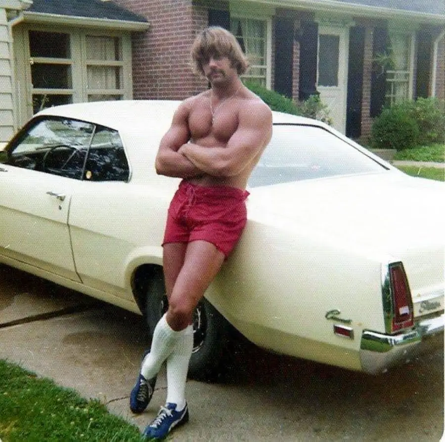 A muscular man in red shorts, long white socks, and blue shoes stands shirtless with arms crossed, leaning against a vintage white car parked in a suburban driveway.
