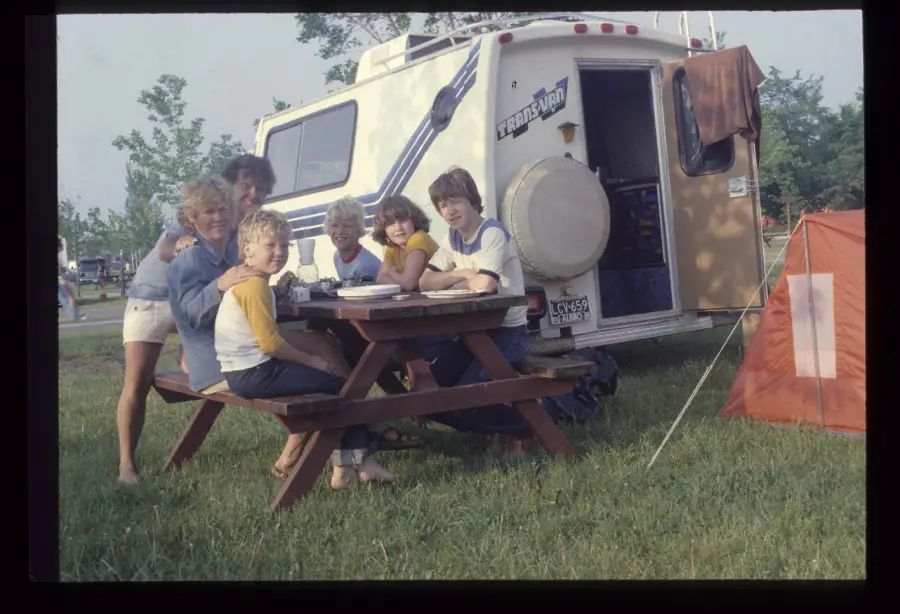 Six people sit at a picnic table beside a camper and an orange tent in a grassy area. They are smiling at the camera. Trees and more campers are visible in the background.