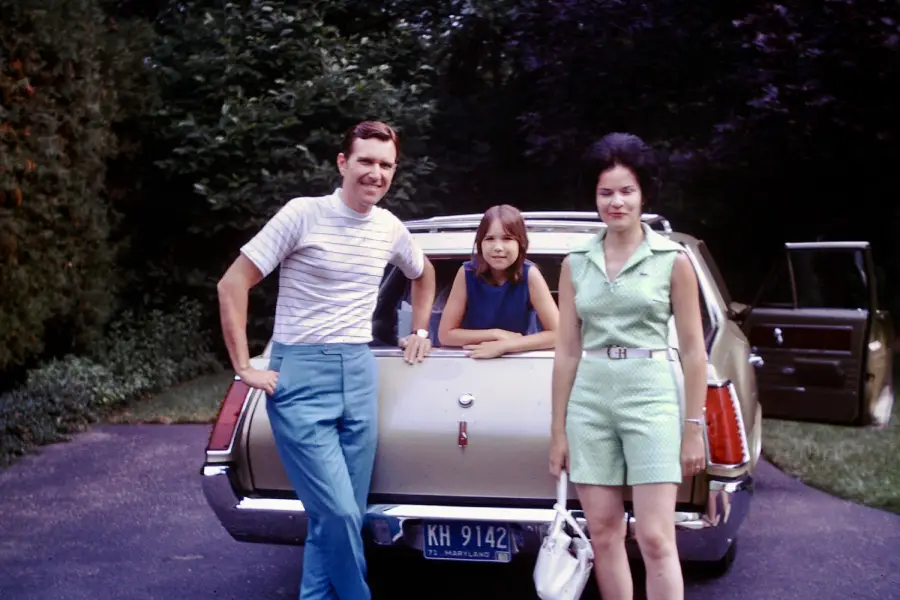 A man, a girl, and a woman stand in front of a beige station wagon parked on a driveway, with trees in the background. The car’s rear door is open, and all three are looking at the camera.