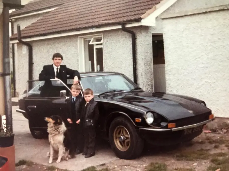 A man, two young boys, and a dog stand by a black classic car parked in front of a light-colored house. The man rests his hand on the car door; everyone is dressed formally.