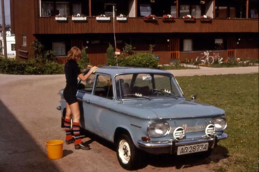 Person in striped socks washes a light blue vintage car with rally lights in front of a brown building, next to a yellow bucket on the ground.