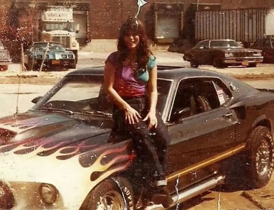 A woman with long hair sits on the hood of a vintage car with flame decals, parked in a lot with other classic cars and industrial buildings in the background. The photo has a faded, retro look.