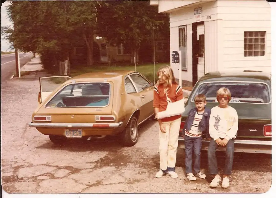 Three kids stand near two parked vintage cars at an old gas station. One child has a cast on her arm, and the other two are sitting on the car bumper. The scene appears to be from the 1970s.