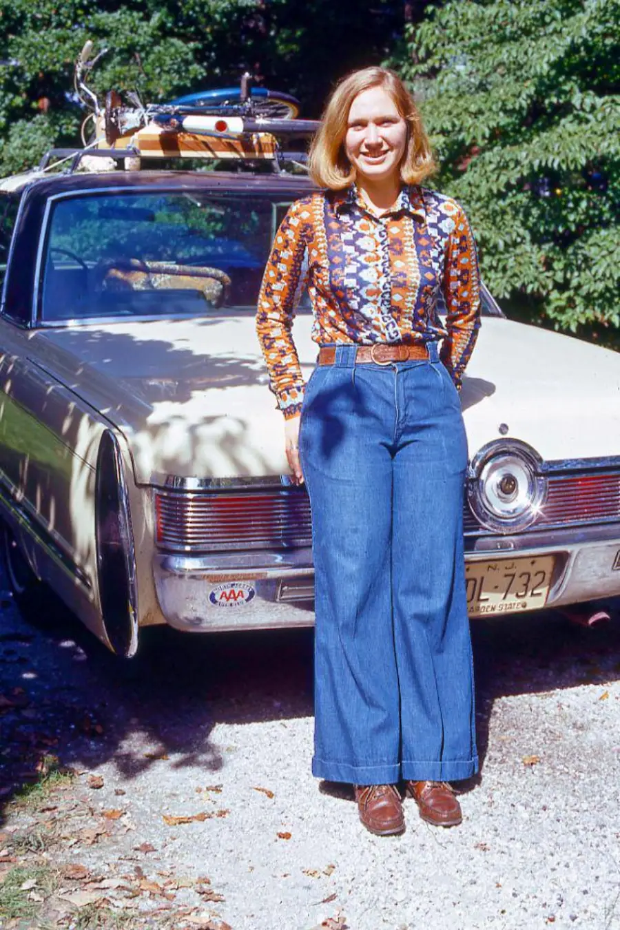 A woman in a patterned shirt and wide-leg jeans stands smiling in front of a vintage car with luggage strapped to the roof, surrounded by greenery on a sunny day.