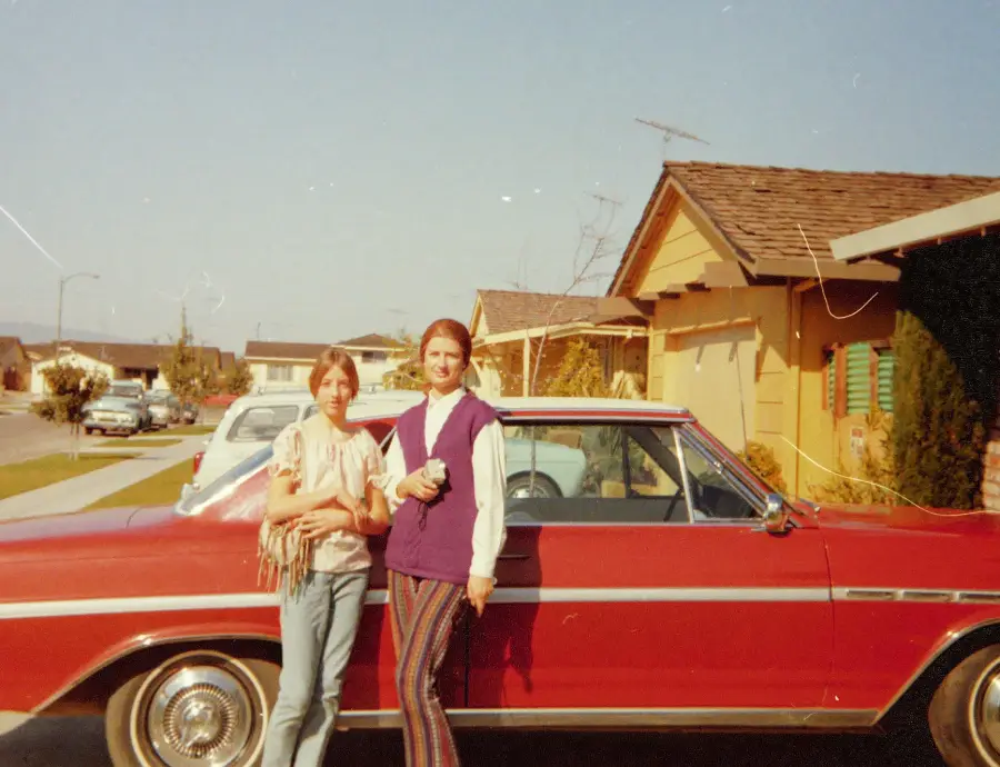 Two women stand in front of a red vintage car parked on a suburban street, with houses and a blue sky in the background.