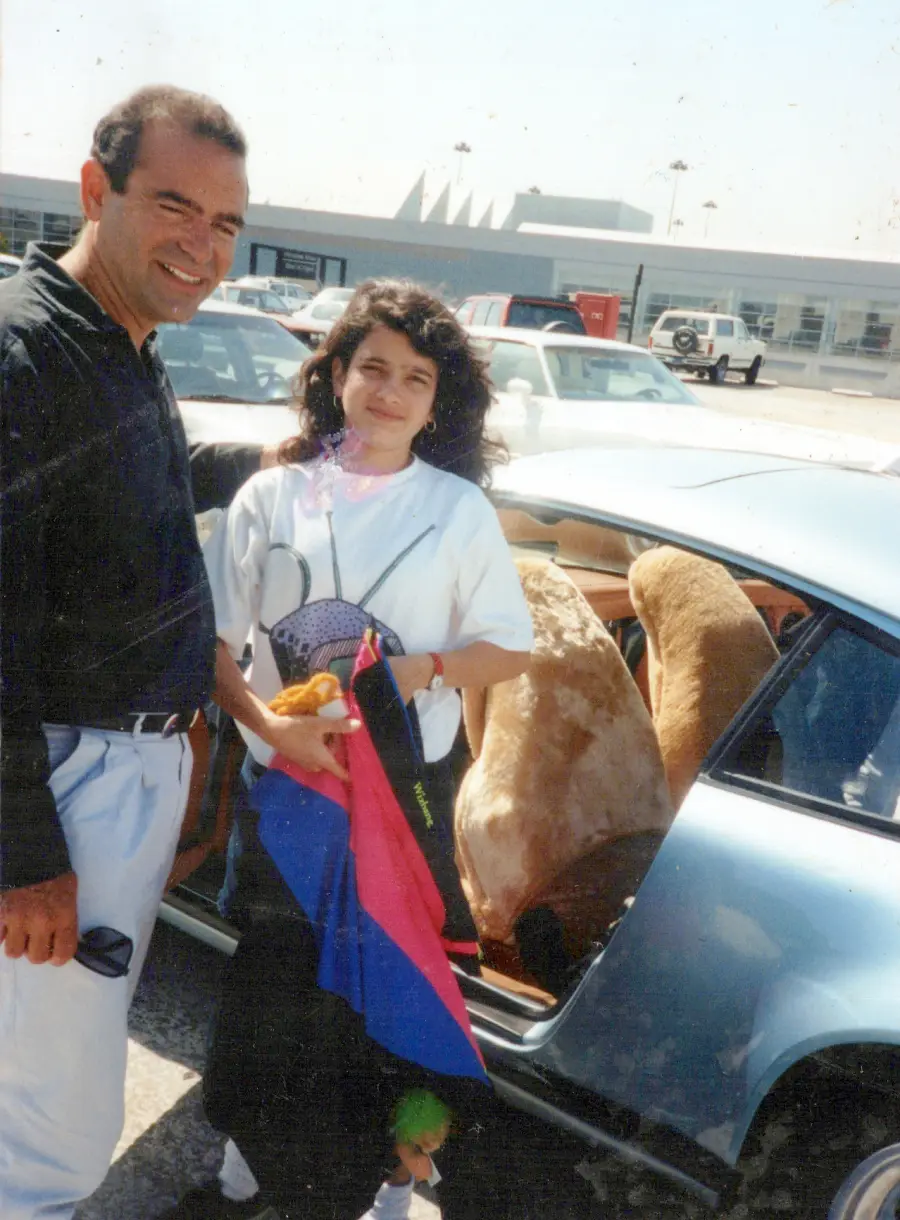 A man and woman stand by a car with two large dogs sitting in the back seat. The woman holds a colorful flag and a stuffed toy. They are in a parking lot on a sunny day.
