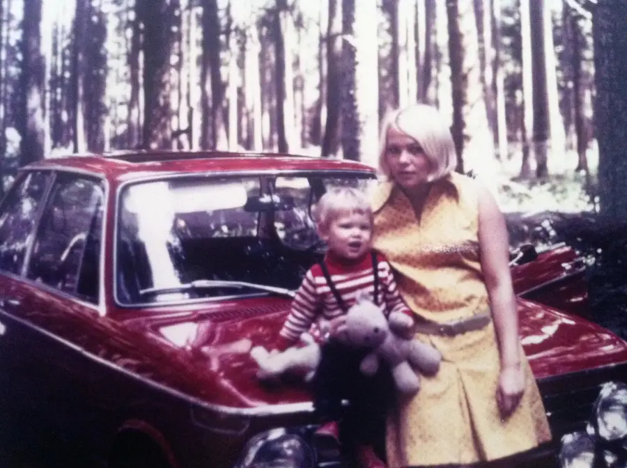 A woman in a yellow dress stands beside a young child holding a stuffed animal, both in front of a red vintage car in a sunlit forest.