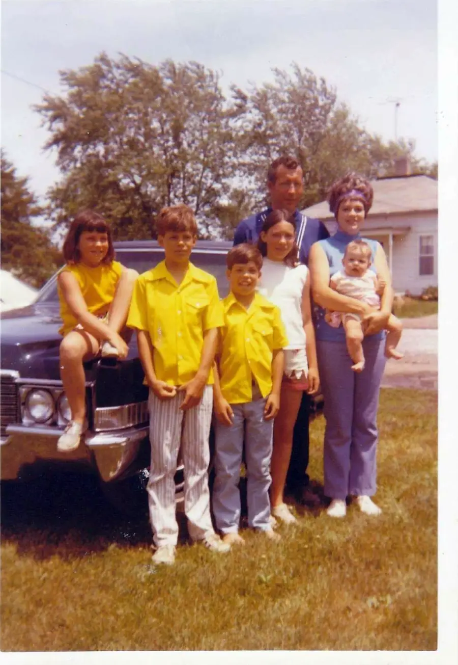 A family of six poses outdoors by a vintage car. Three children wear yellow tops, a woman holds a baby, and a man stands behind them. There’s a house and trees in the background on a sunny day.