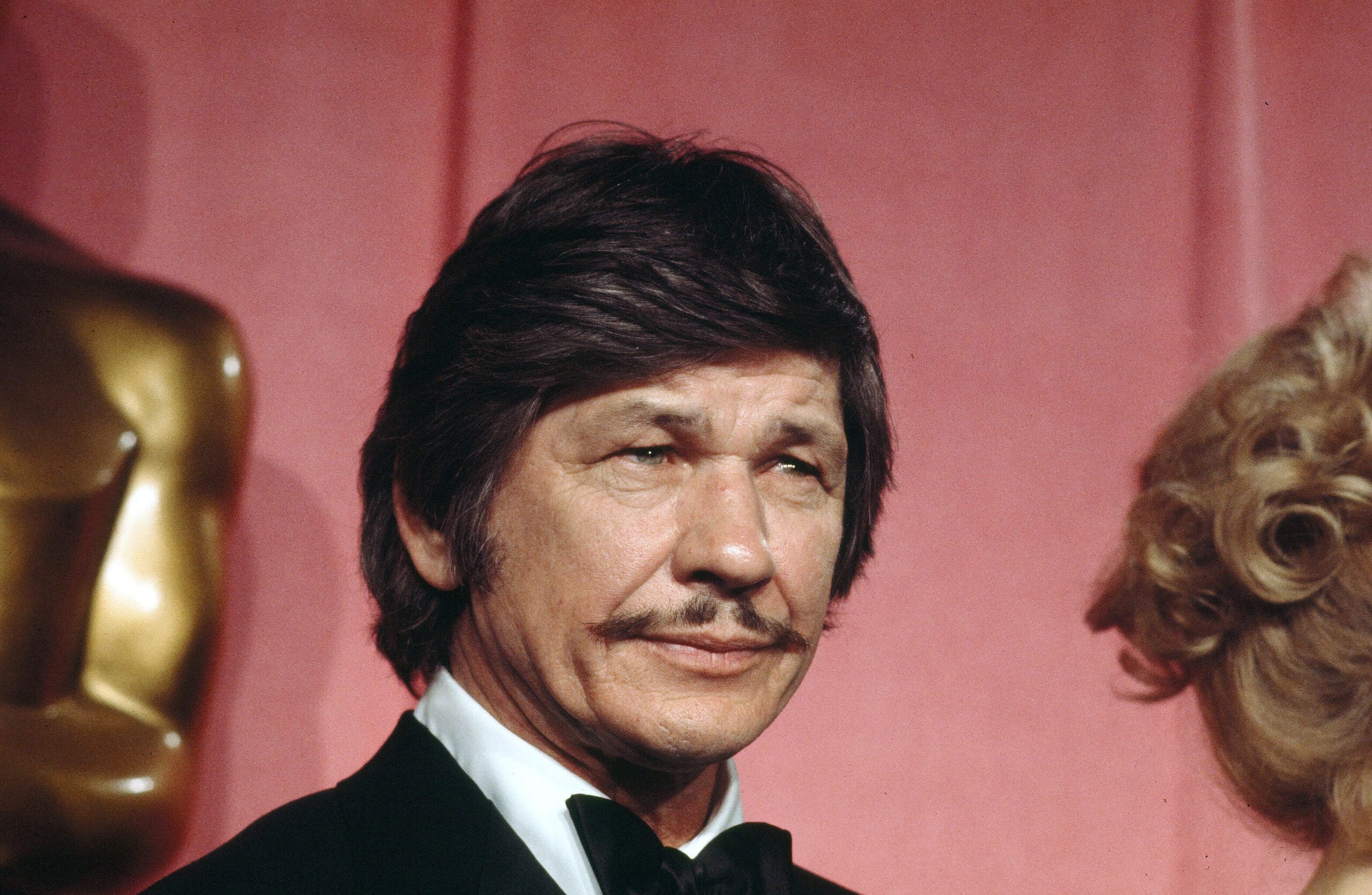 A man with dark hair and a mustache wearing a tuxedo stands in front of a pink backdrop and a gold statue. A woman’s blonde hair is partially visible on the right side of the image.