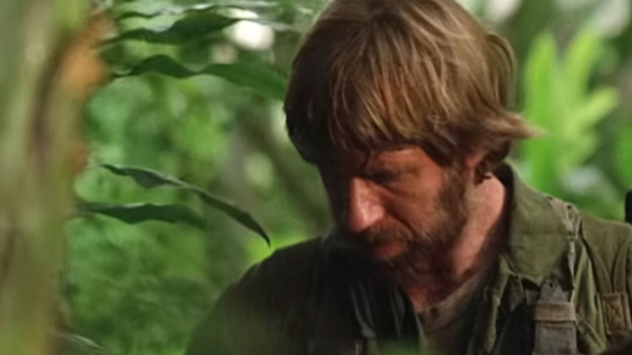 A man with a beard and shaggy hair, dressed in green clothing, stands in dense jungle foliage, looking down with a serious expression.