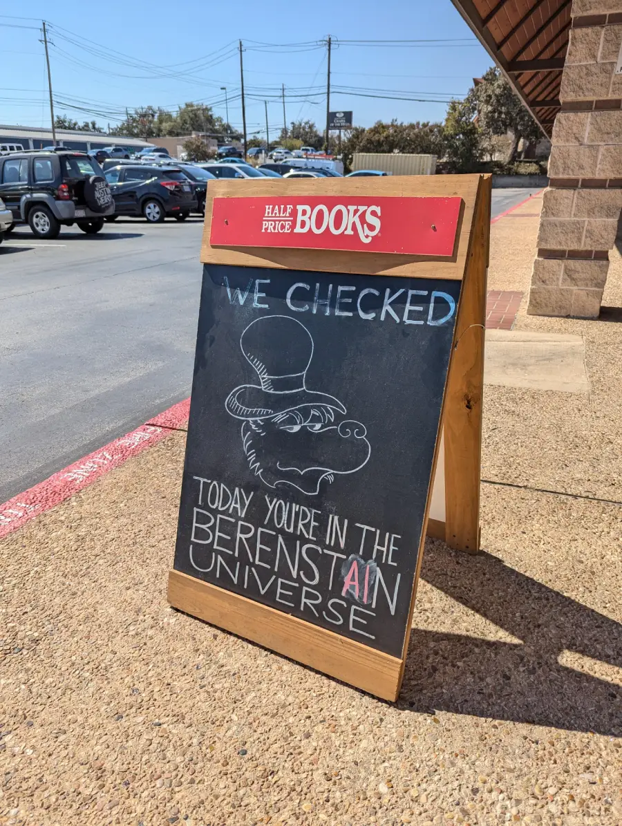 A chalkboard sign outside Half Price Books reads, “WE CHECKED. TODAY YOU’RE IN THE BERENSTAIN UNIVERSE,” with a drawing of a bear wearing a hat. Cars and a building are visible in the background.