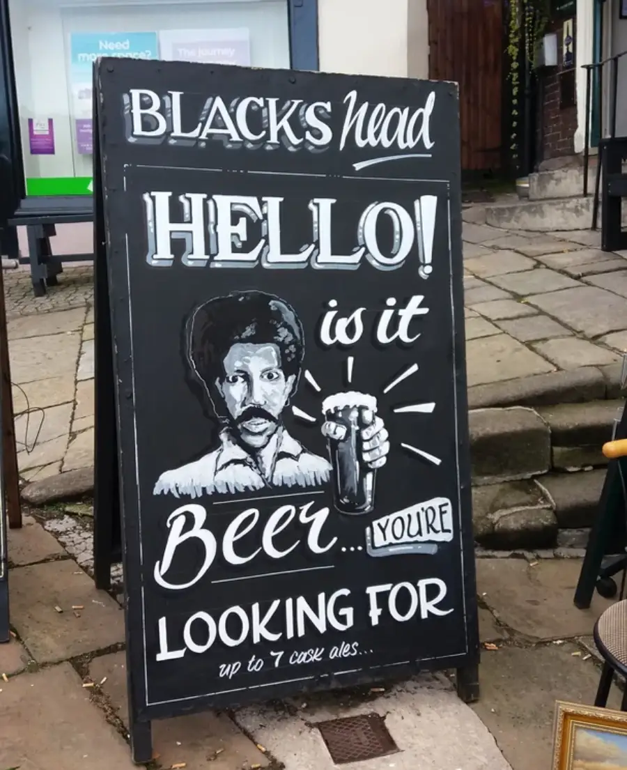 A chalkboard sign outside a pub shows a hand-drawn man holding a beer with the text: “BLACKS head. HELLO! Is it beer you’re looking for. Up to 7 cask ales.”