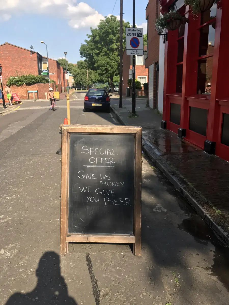 A chalkboard sign on a sidewalk reads: “SPECIAL OFFER: GIVE US MONEY, WE GIVE YOU BEER.” The sign is in front of a red building on a quiet street with a few cars and people in the background.