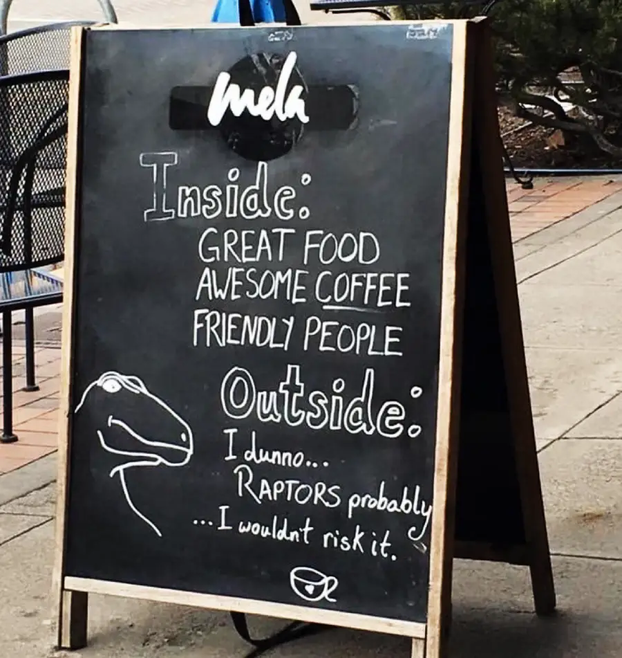 A chalkboard sign outside a cafe reads: “Inside: Great food, awesome coffee, friendly people. Outside: I dunno... raptors probably... I wouldn’t risk it,” with a cartoon dinosaur drawing.