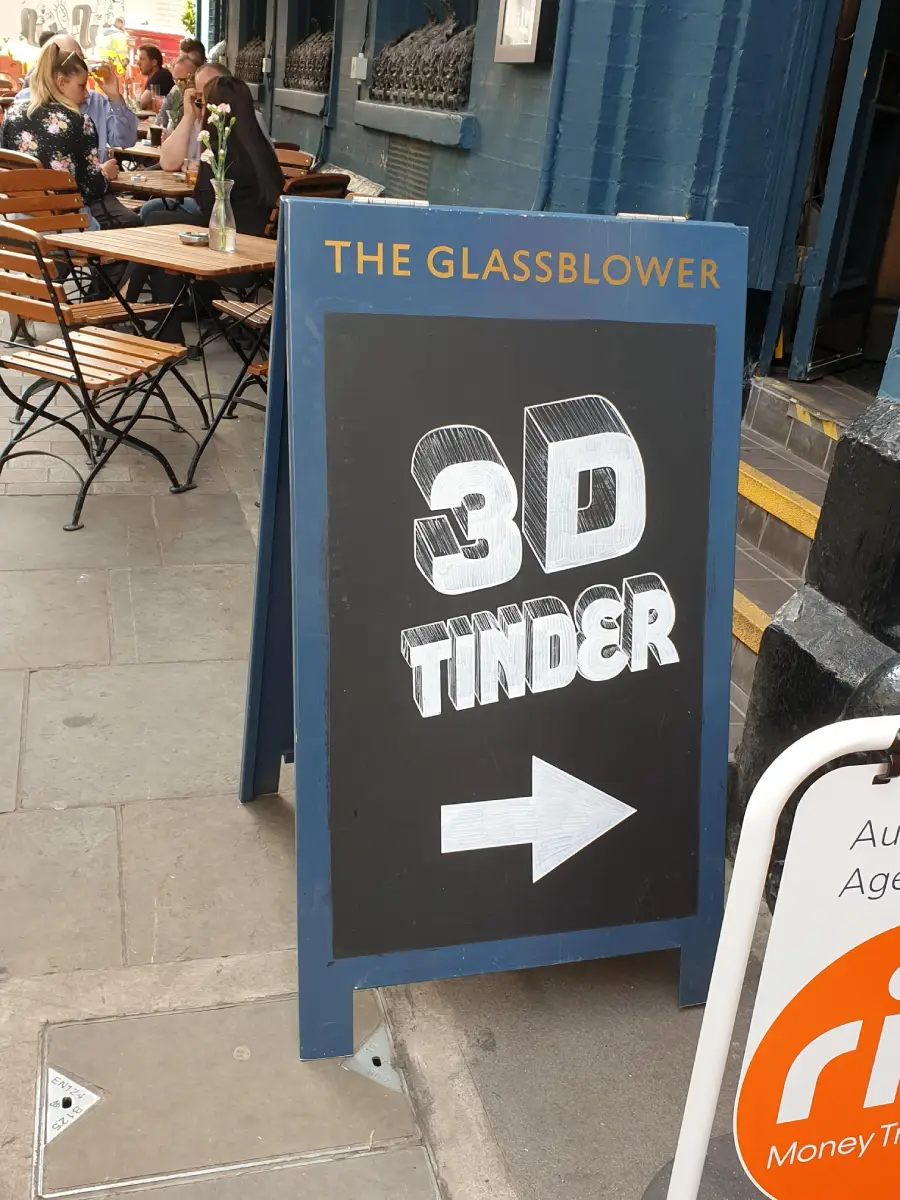 A chalkboard sign outside a café reads “3D Tinder” with an arrow, humorously referring to in-person dating; people are sitting at tables in the background.
