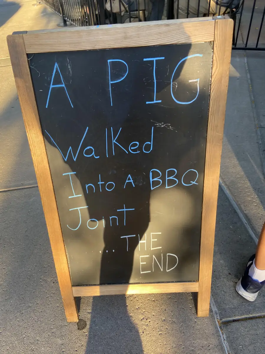 A chalkboard sign reads: “A pig walked into a BBQ joint... THE END.” The sign stands on a sidewalk with sunlight casting shadows.