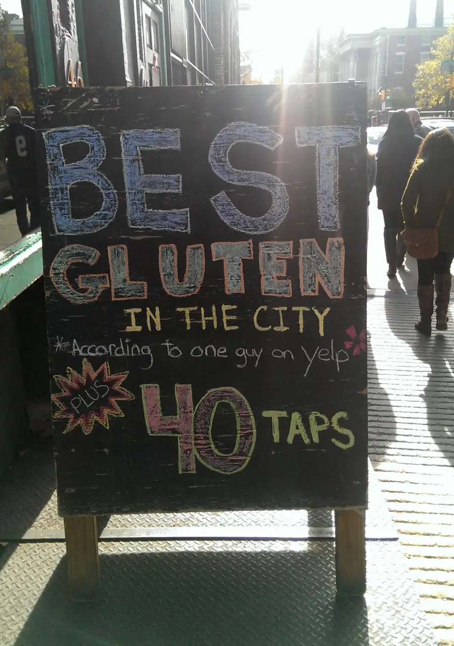 A sidewalk chalkboard sign reads: “BEST GLUTEN IN THE CITY *According to one guy on Yelp* plus 40 TAPS” in colorful, bold letters. People walk on the sidewalk in sunlight behind it.