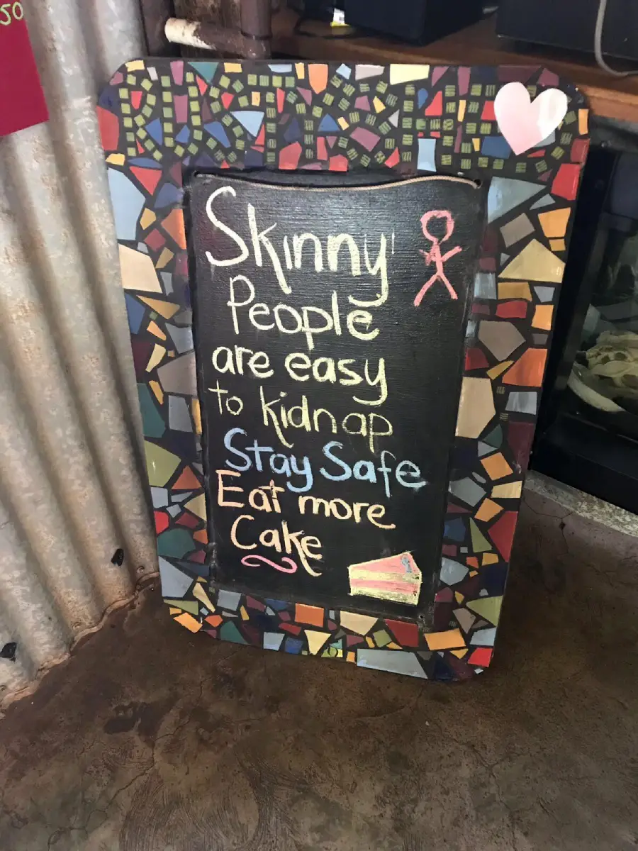 A colorful chalkboard sign reads: “Skinny people are easy to kidnap. Stay safe. Eat more cake,” with a stick figure and a drawing of a slice of cake at the bottom.