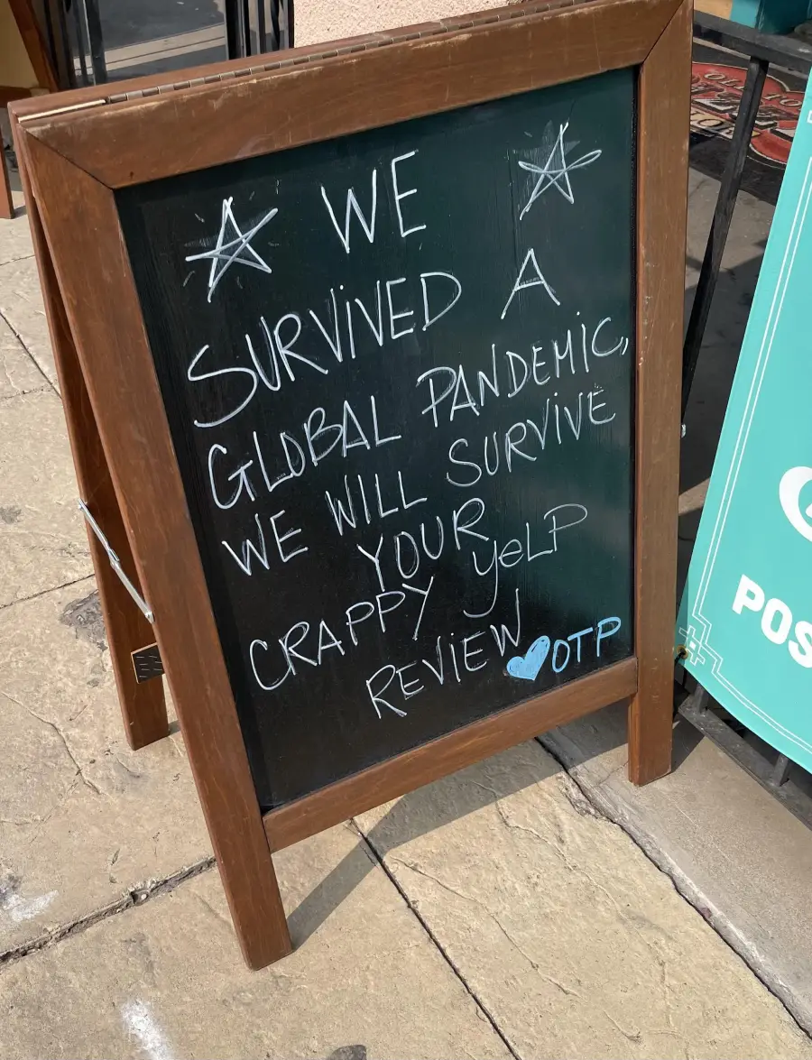 A chalkboard sign on a sidewalk reads: “We survived a global pandemic, we will survive your crappy Yelp review. OTP” with two drawn stars and a blue heart.