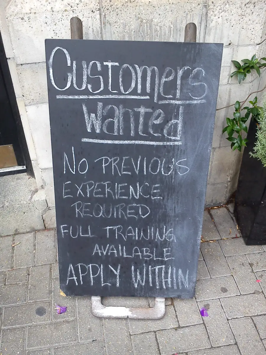 A chalkboard sign reads: “Customers Wanted. No previous experience required. Full training available. Apply within.” It’s placed on a sidewalk near a building entrance.