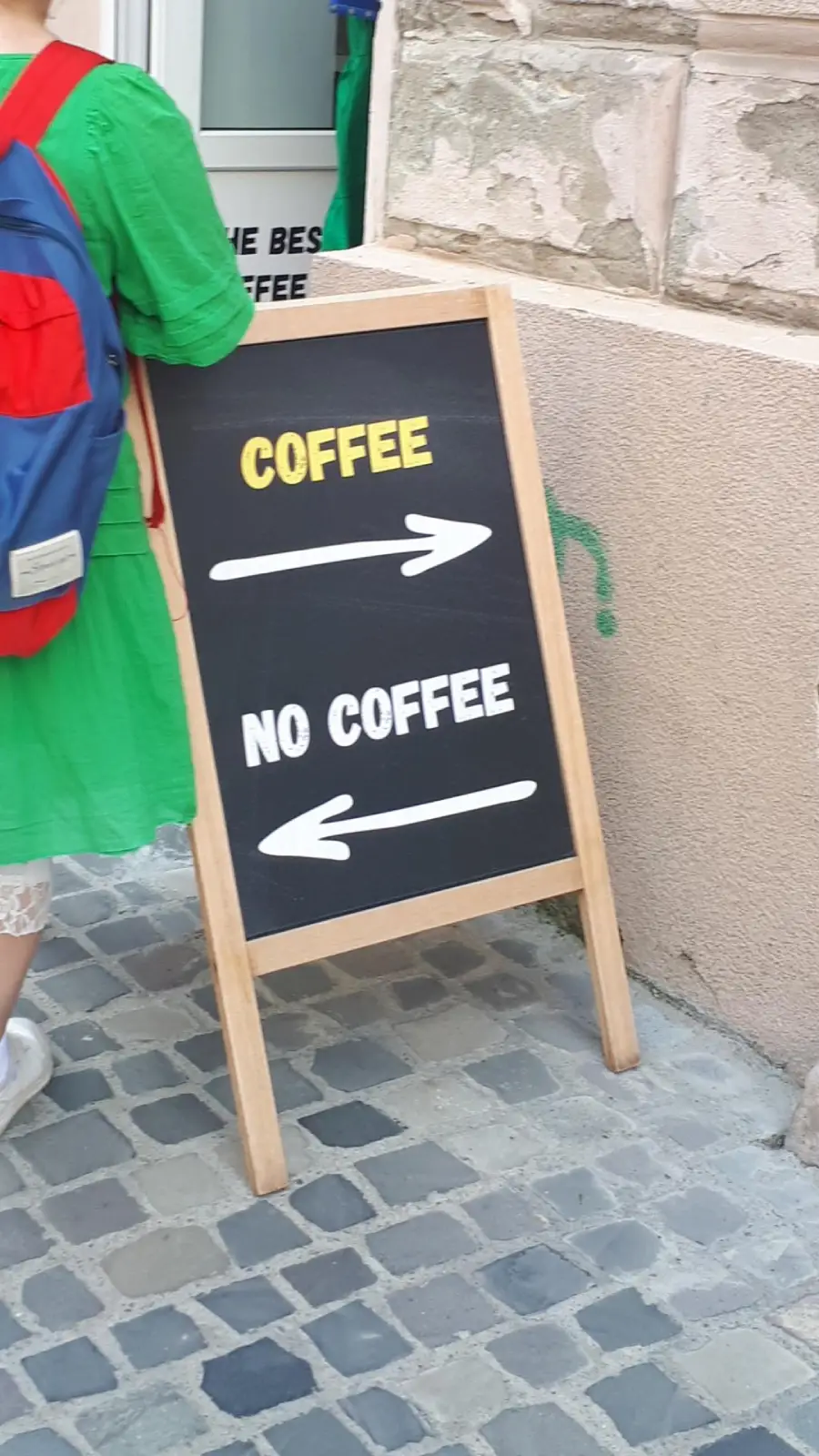 A chalkboard sign on a cobblestone street points right for “COFFEE” and left for “NO COFFEE.” A person in a green dress and red-blue backpack stands nearby.