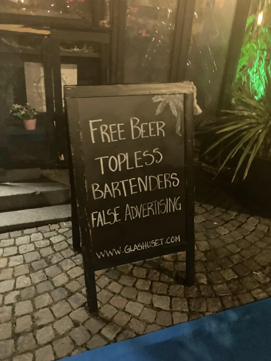 A chalkboard sign on a cobblestone sidewalk reads: “Free Beer, Topless Bartenders, False Advertising” with a website below. Plants and a glass door are in the background.