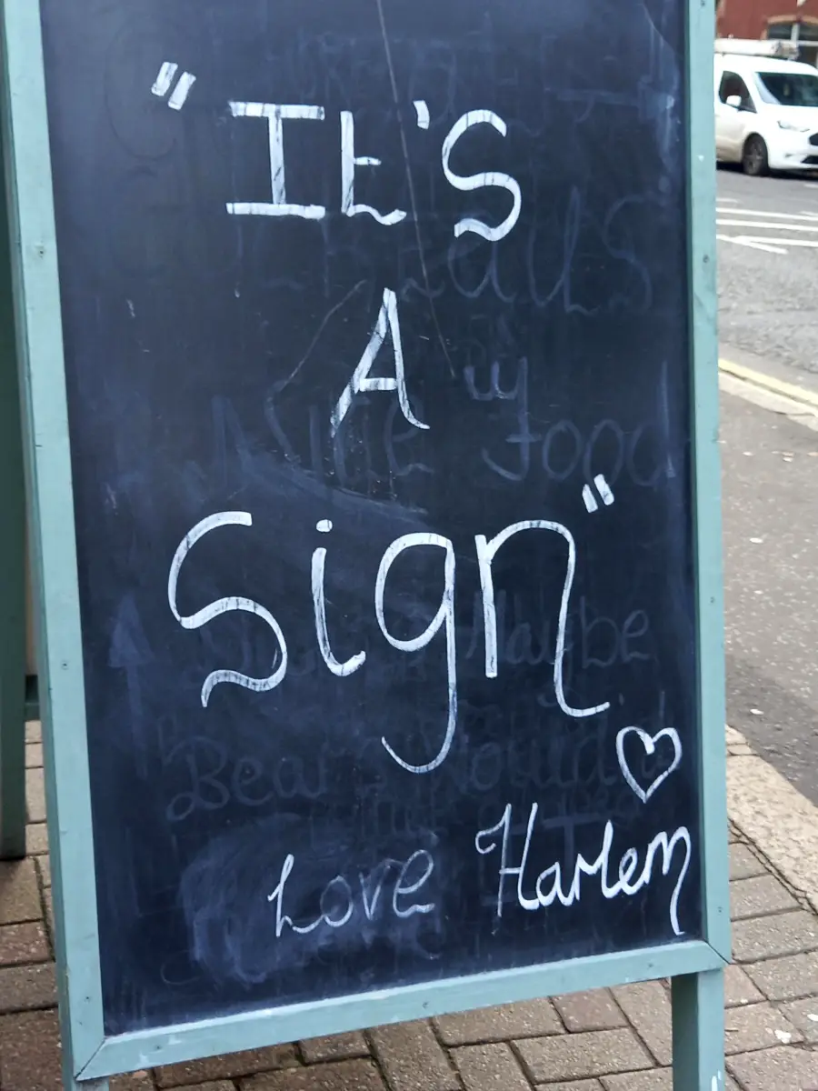 A chalkboard sidewalk sign reads: “It’s a sign” with a heart and “Love Harlem” written at the bottom. The sign is on a sidewalk near a street.
