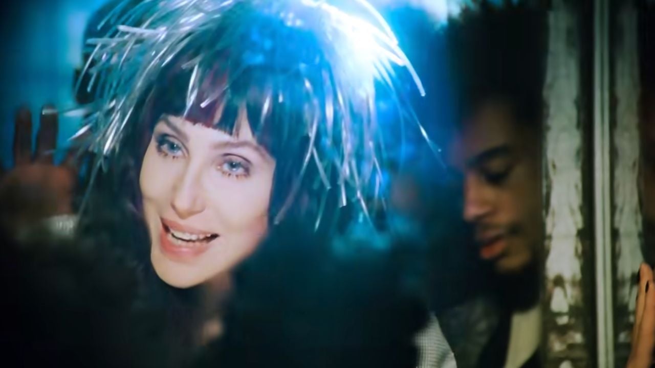 A woman with blue eyes and a shiny, silver headpiece smiles brightly in the foreground, while a man with closed eyes stands beside her under blue light.