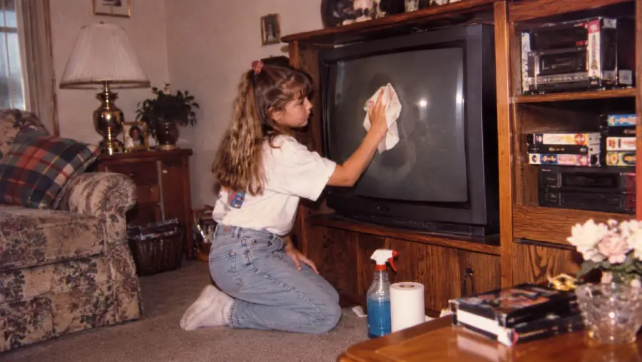 A girl kneels on the floor cleaning a large box-style TV with a cloth. Cleaning supplies are nearby. The room has a sofa, lamp, and shelves with VHS tapes.