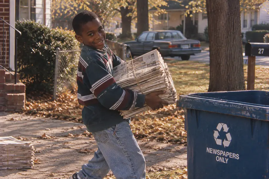 Smiling boy in striped sweater carries a stack of newspapers to a blue recycling bin labeled “Newspapers Only” on a leafy suburban street.