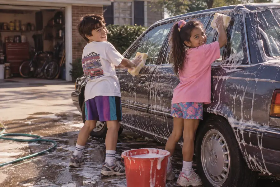 Two children wearing colorful clothes wash a soapy car together outdoors. A red bucket and sponge are in the foreground, and an open garage with bikes is visible in the background.